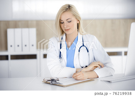 Woman-doctor at work while sitting at the desk in hospital or clinic. Blonde cheerful physician filling up medication history record form 76219938