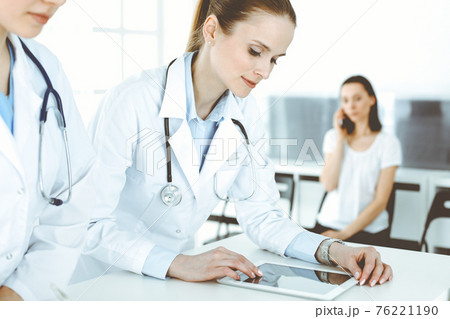 Two woman-doctors at work with patient at background. Female physicians filling up medical documents or prescription while standing in hospital reception desk. Data in medicine 76221190