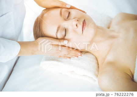 Happy woman enjoying facial massage with closed eyes in spa salon. Relaxing treatment in medicine and Beauty concept 76222276