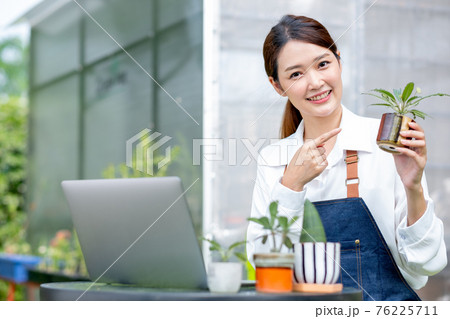 Beautiful Asian girl hold pot of plant and look at camera also pointing to pot stay with laptop Beautiful Asian girl hold pot of plant and look at camera also pointing to pot stay with laptop 76225711