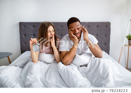 Interracial married couple having problem with waking up in morning, sitting sleepy in bed with alarm clock Interracial married couple having problem with waking up in morning, sitting sleepy in bed with alarm clock 76228223