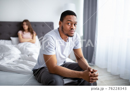 Erectile dysfunction, sex or relationship problems. Upset black guy sitting on bed, his dissatisfied wife on background 76228343