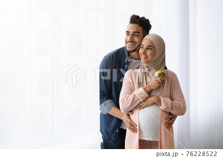 Portrait Of Happy Pregnant Islamic Couple Standing Near Window At Home Portrait Of Happy Pregnant Islamic Couple Standing Near Window At Home 76228522