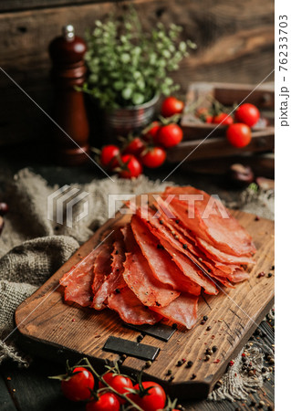 Delicious smoked sausage on a dark wooden board with tomatoes and spices. Delicious handmade snack made from natural ingredients. Pieces of dry pork, beef, chicken snacks. Meat products. Close-up. 76233703