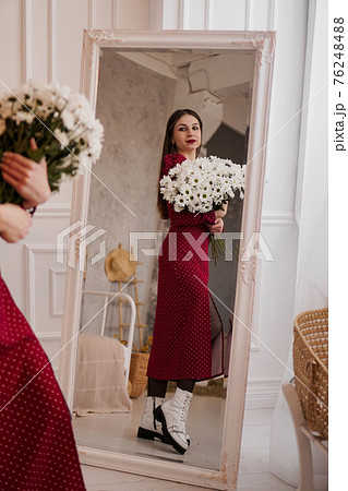 Beautiful brunette in a red dress with a bouquet of daisies looks in the mirror at home 76248488