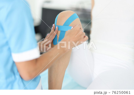 Rehabilitation therapist gluing blue tapes on woman knee closeup Rehabilitation therapist gluing blue tapes on woman knee closeup 76250061