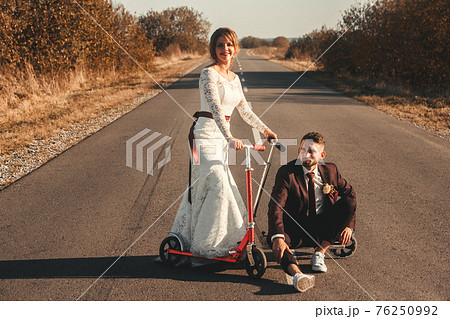 Smiling wedding couple riding a on scooters along the road outside the city at sunset. Smiling wedding couple riding a on scooters along the road outside the city at sunset. 76250992