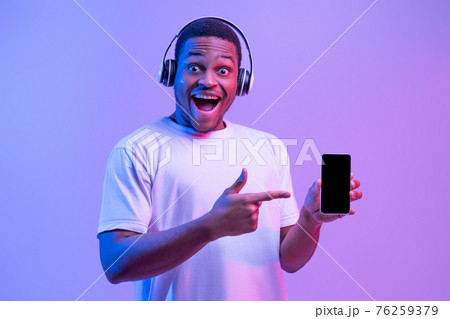 Online Music Platform. Excited African Man In Headphones Pointing At Black Smartphone 76259379