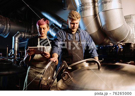 Young woman with digital tablet standing next to male colleague opening lid of huge steel cistern Young woman with digital tablet standing next to male colleague opening lid of huge steel cistern 76265000