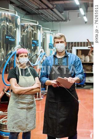 Young brewers in workwear and protective masks standing in front of camera against huge steel tanks Young brewers in workwear and protective masks standing in front of camera against huge steel tanks 76265011