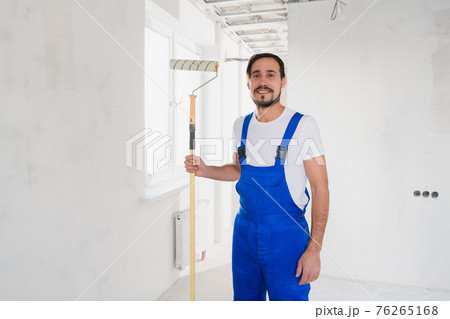 A handyman builder in a blue overalls holds a roller in his hand and poses at the camera A handyman builder in a blue overalls holds a roller in his hand and poses at the camera 76265168