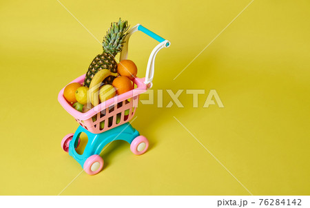 Toy kids pink shopping cart full of assorted exotic fruits isolated on yellow background , copy space. Summer concepts 76284142