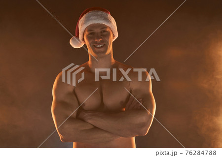 Portrait of muscular man wearing Christmas Santa hat, folded hands on smoky background 76284788