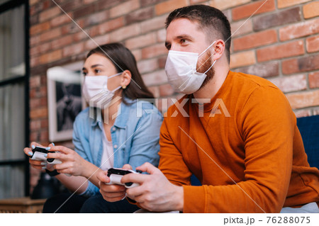 Close-up portrait of happy young couple wearing medical protective masks playing video games on console 76288075