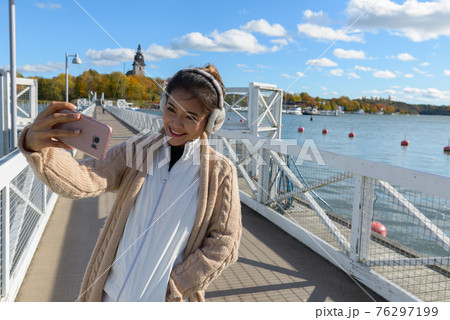 Young happy Asian woman smiling while taking selfie picture with mobile phone on the pier 76297199