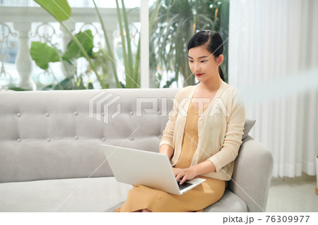 Surfing web at home. Side view of attractive young asian woman working on laptop and smiling while sitting on the couch at home 76309977