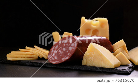 Several types of cheese and piece of salami loaf are on black stone board. 76315670