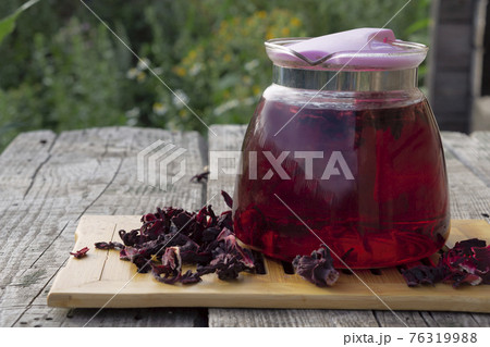 Glass pot of red herbal Hibiscus tea with ingredients for tea against a blurred green meadow Glass pot of red herbal Hibiscus tea with ingredients for tea against a blurred green meadow 76319988