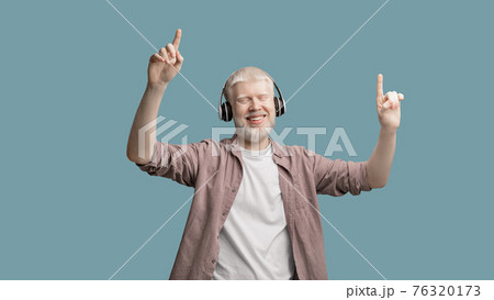 Happy albino man enjoying music in wireless headphones, dancing and raising fingers up over turquoise studio background Happy albino man enjoying music in wireless headphones, dancing and raising fingers up over turquoise studio background 76320173