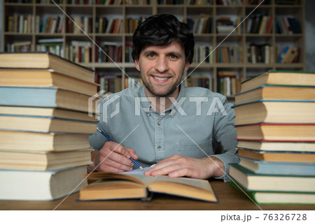 Man student sits at a table in the library, littered with books. Man student sits at a table in the library, littered with books. 76326728