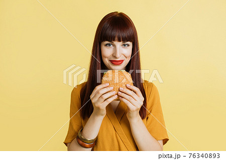 Junk and fast food concept. Cheerful young pretty brught woman in yellow shirt and red lipstick, holding fresh tasty burger, standing over yellow background. Studio Shot with Copy Space 76340893