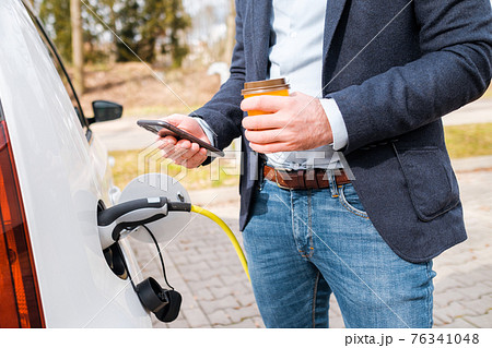 Business man standing near the charging electric car , drinking coffee and using his smartphone. Zero emission car.  76341048
