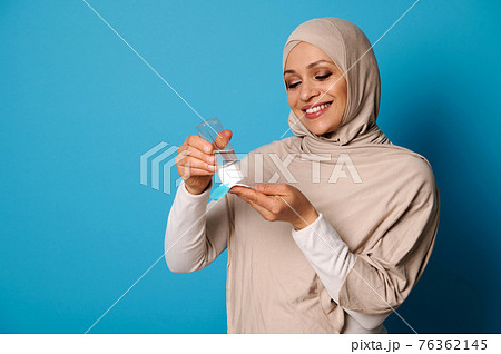 Smiling muslim woman wearing beige hijab applying micellar makeup lotion to cotton pad. Blue background, copy space 76362145
