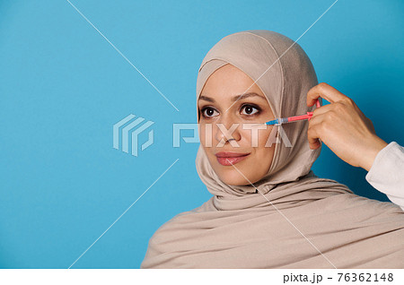 Attractive young Muslim woman with perfect makeup and clean healthy skin, posing with a syringe near her face over blue background 76362148