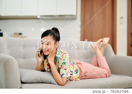 Slender young woman relaxing at home on a sofa chatting on her mobile phone with a happy smile 76362566