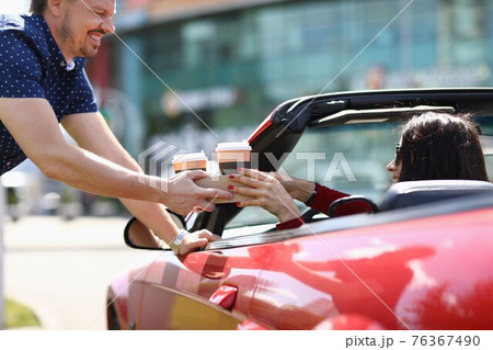 Man serving two glasses of coffee to woman in red convertible 76367490
