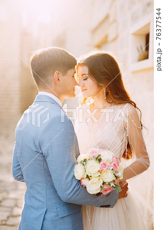 The bride and groom hug on a beautiful old street of Perast near a white stone wall, close-up The bride and groom hug on a beautiful old street of Perast near a white stone wall, close-up 76377544
