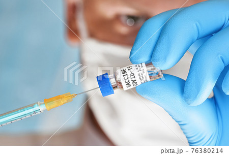 Man hand in blue glove hold small vial with coronavirus vaccine (label own design, not real product), to inject it with green orange syringe, blurred face with white virus mask in background Man hand in blue glove hold small vial with coronavirus vaccine (label own design, not real product), to inject it with green orange syringe, blurred face with white virus mask in background 76380214