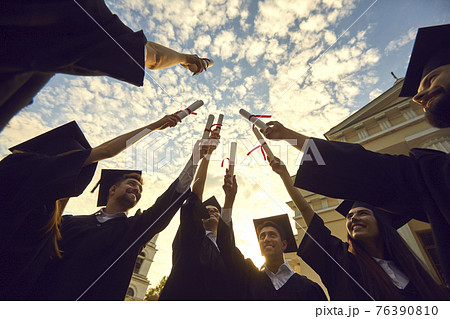 Bottom view graduates lift their diplomas and stack them together against a blue sky with clouds. 76390810