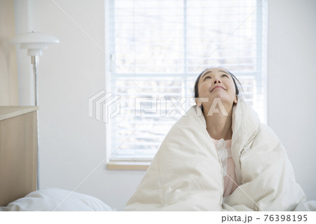 Asian woman wrapping herself with blanket while sitting on bed 76398195