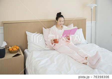 Asian woman with hair bun in pink pajamas on bed reading book and drinking coffee Asian woman with hair bun in pink pajamas on bed reading book and drinking coffee 76398306