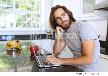 Caucasian man sitting in kitchen using laptop and thinking Caucasian man sitting in kitchen using laptop and thinking 76401300