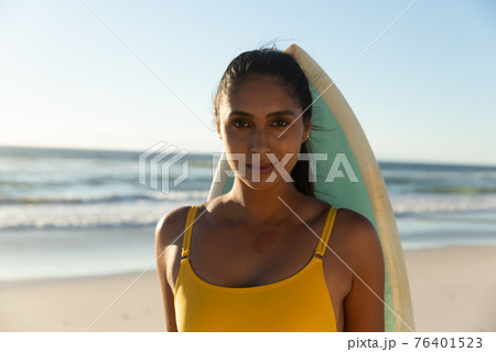 Portrait of mixed race woman with surfboard on the beach looking to camera 76401523
