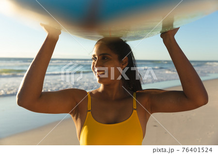 Happy mixed race woman on the beach carrying surfboard on head 76401524