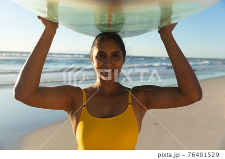 Happy mixed race woman on the beach carrying surfboard on head Happy mixed race woman on the beach carrying surfboard on head 76401529