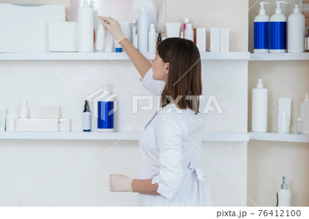 Cosmetologist in a white coat taking beauty products from the shelf 76412100