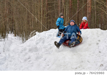 Grandfather and his grandchildren ride on a snow slide on a rubber circle Grandfather and his grandchildren ride on a snow slide on a rubber circle 76420960