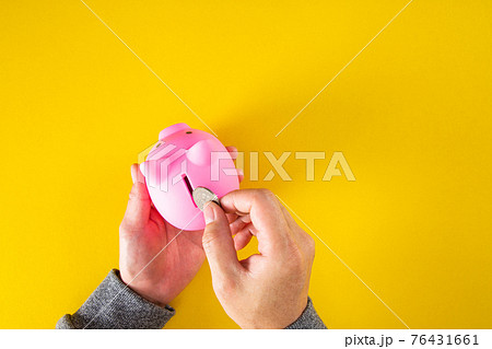 Hand puts a coin into the piggy bank coin on a yellow background. Top view Hand puts a coin into the piggy bank coin on a yellow background. Top view 76431661