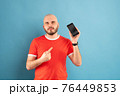 A bald middle-aged man with a beard and in a red T-shirt on a blue background. Holds a phone in his hand and points at it with the finger of his other hand 76449853