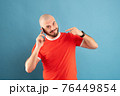 A bald middle-aged man with a beard and in a red T-shirt on a blue background. Holds a phone in his hand and points at it with the finger of his other hand 76449854