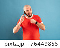 A bald middle-aged man with a beard and in a red T-shirt on a blue background. Holds a phone in his hand and points at it with the finger of his other hand 76449855