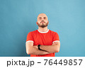 A middle-aged man with a beard in a red T-shirt on a blue background stands with his arms outstretched and rejoices in victory. Isolated. 76449857
