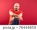 A middle-aged man with a beard in a red T-shirt on a red background joyfully points to the right. Isolated. 76449859