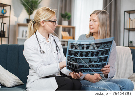 Middle aged attractive high-skilled caring female doctor explaining results of x-ray MRI image to her senior woman patient during visiting her at home. Computed tomography 76454893