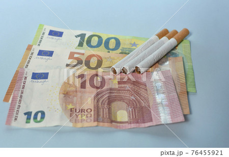 Cigarettes and money on blue background. Euro banknotes and coins, cost of smoking, cigarette price concept. Selective focus Cigarettes and money on blue background. Euro banknotes and coins, cost of smoking, cigarette price concept. Selective focus 76455921