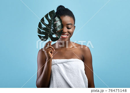 Pretty black woman with smooth skin wrapped in towel hiding behind tropical plant on blue studio background 76475148